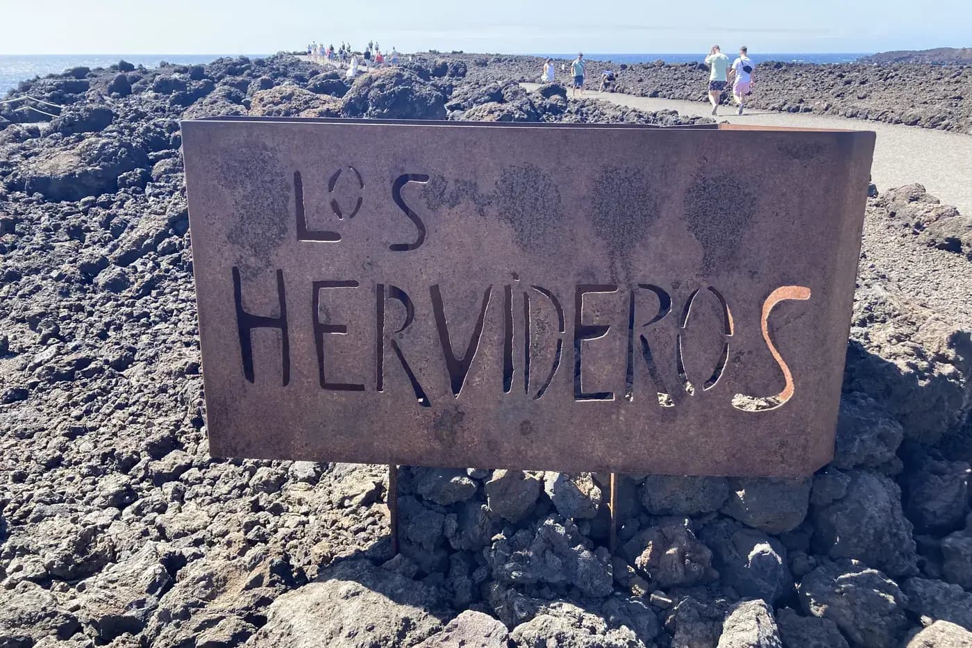 Schild „Los Hervideros“ im Vulkangestein, mit Besuchern entlang der Küste